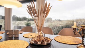 Une table à manger dressée avec des sets de table en fibres tressées, un vase en verre contenant des roseaux séchés et un plateau en métal rempli de grains de café.