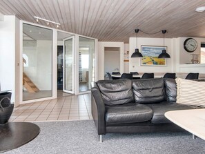 A modern living room with a black leather sofa, a dining area with a table and chairs, and a kitchen area with a clock and pendant lights. - 4 Star Holiday Home in Harboore (Harboore)