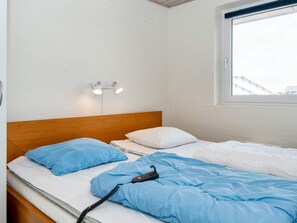 A single bed with a wooden headboard, white bedding, and a blue pillow. There is a bedside table with a lamp and a power outlet. A window with a view of buildings is visible. - 4 Star Holiday Home in Harboore (Harboore)