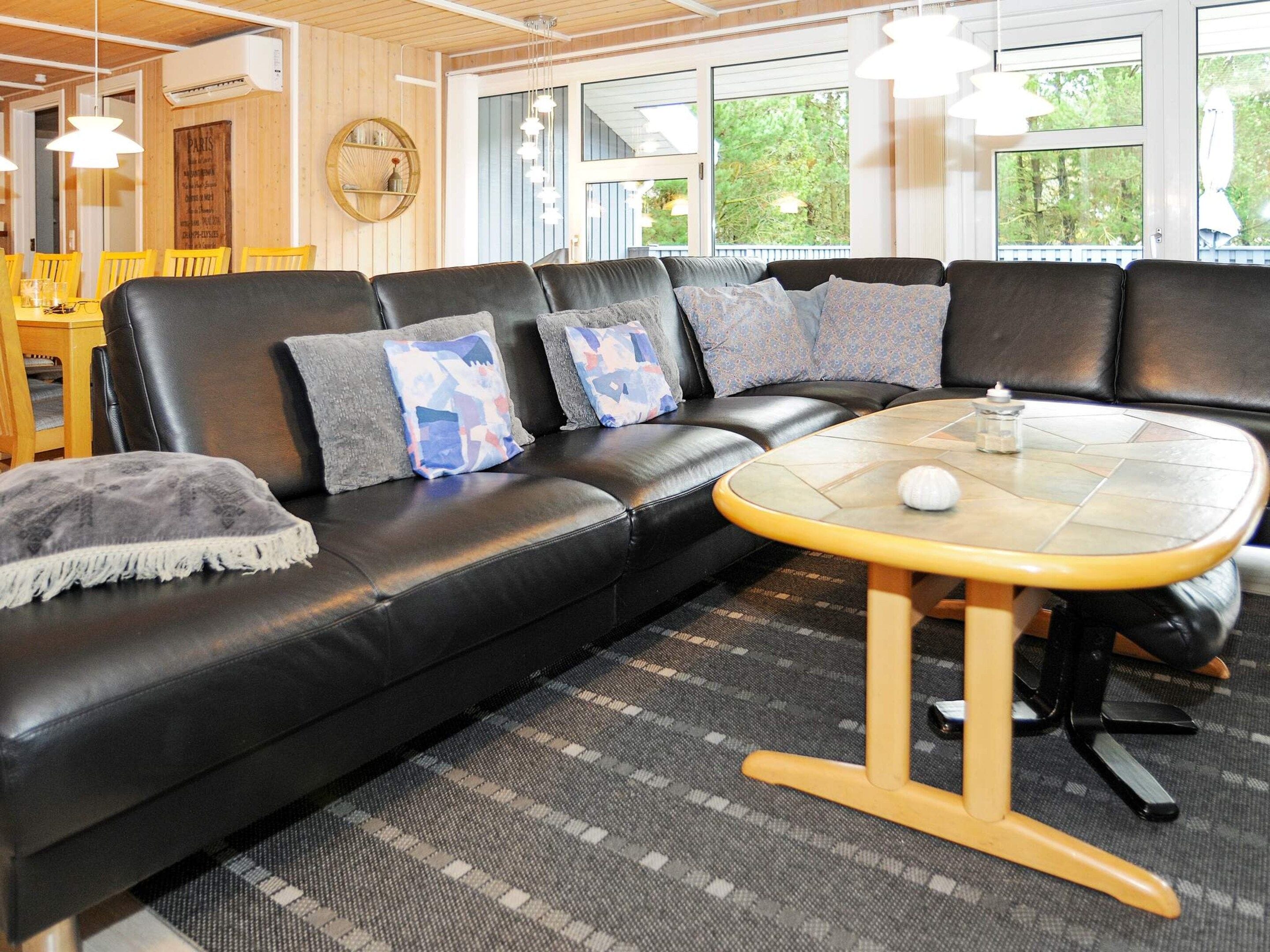 A modern living room with a black leather sofa, patterned cushions, a small round table, and a view of greenery outside.