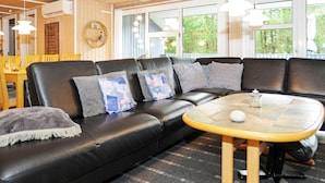 A modern living room with a black leather sofa, patterned cushions, a small round table, and large windows with a view of greenery. - 4 Star Holiday Home in Norre Nebel (Norre Nebel)