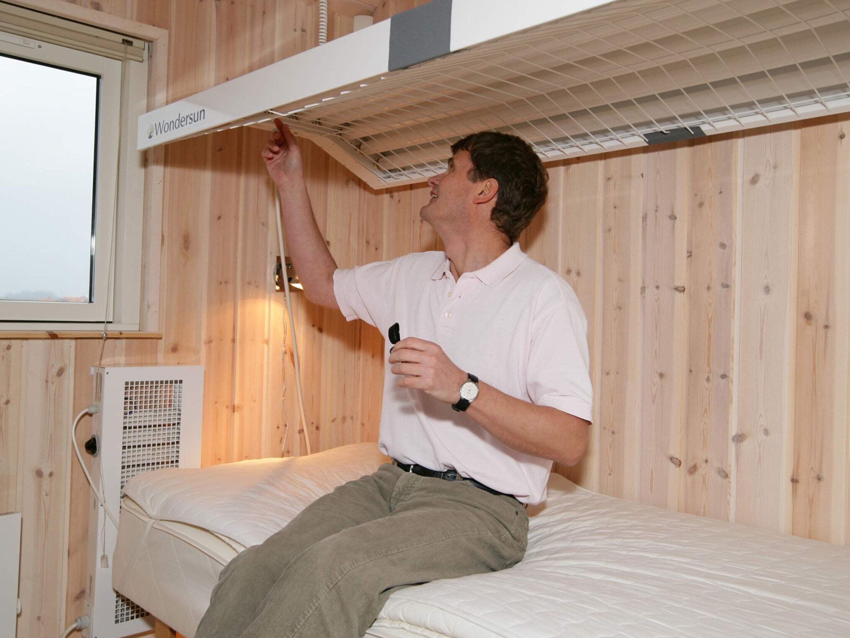 Eine Person sitzt auf einem Bett in einem Zimmer mit Holzwänden, einem Fenster und einem Deckenventilator.