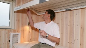 A person sitting on a bed in a room with wooden walls, a window, and a ceiling fan.