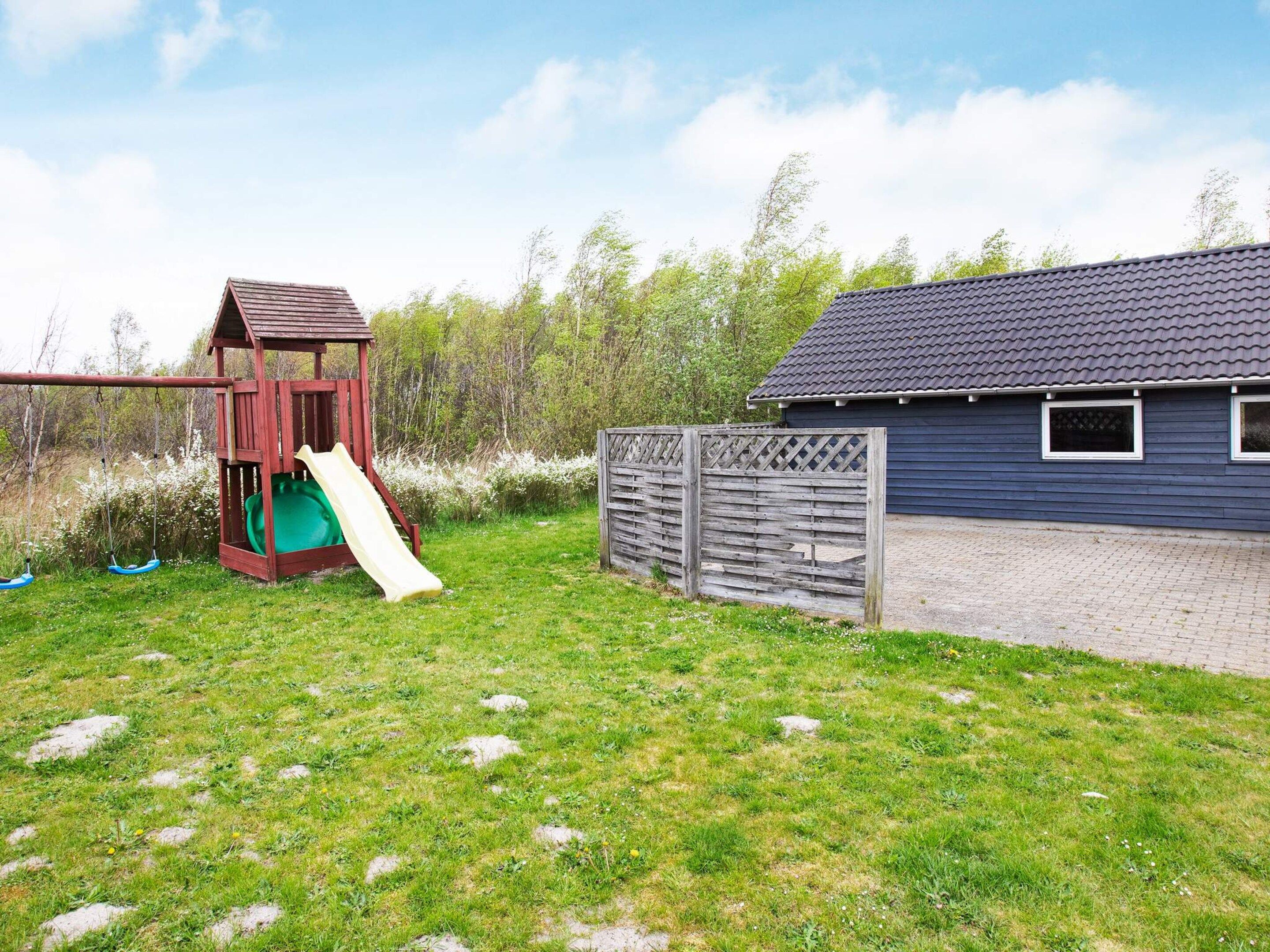 Ein Spielplatz mit Rutsche und Schaukeln, einem Holzzaun und einem Gebäude mit dunklem Dach.
