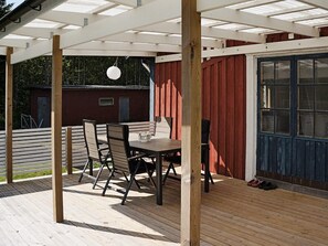 An outdoor patio with a wooden table and chairs, a pergola overhead, and a red building in the background.