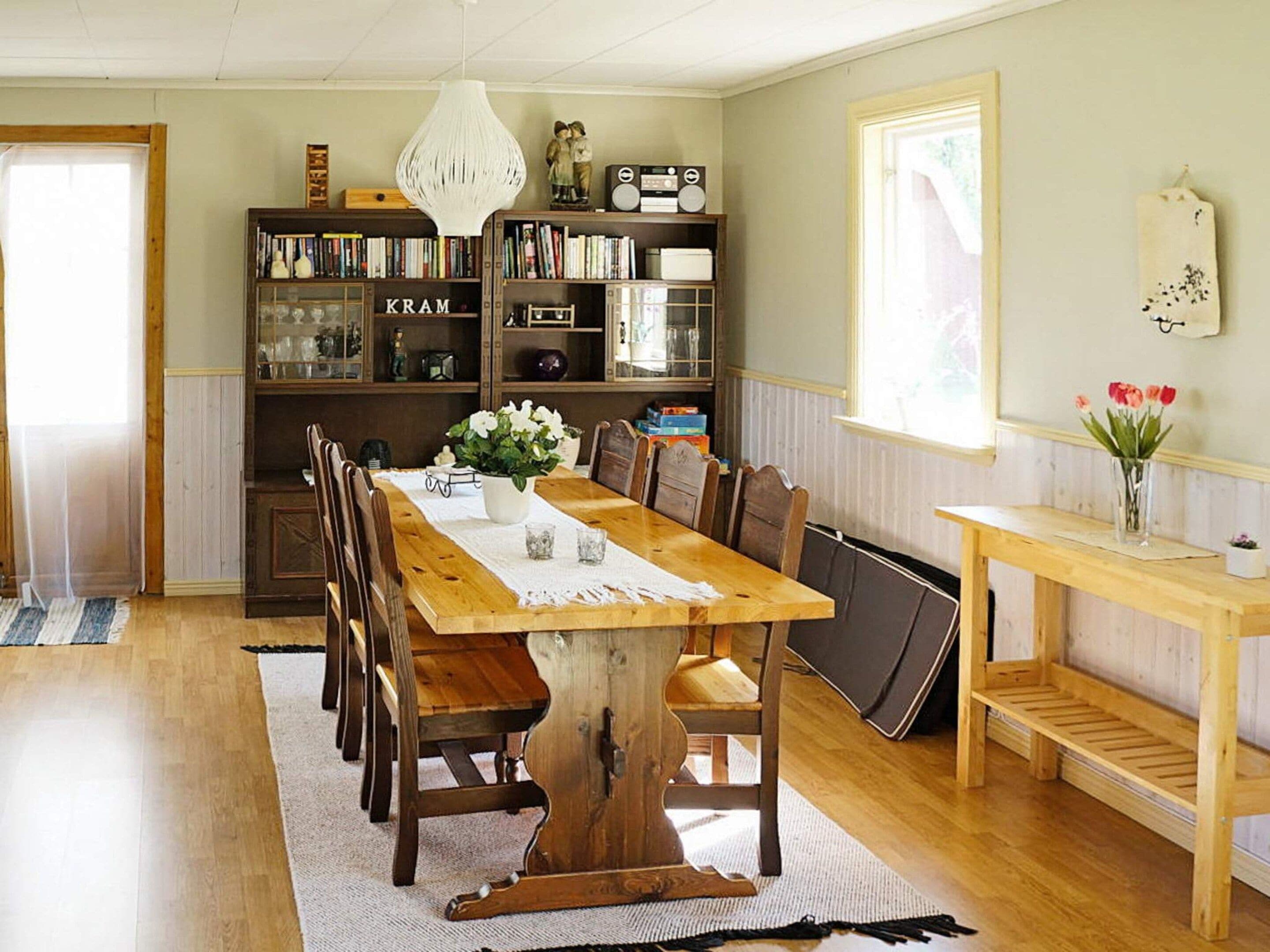 A dining area with a wooden table, chairs, a chandelier, bookshelves, and a window with flowers.