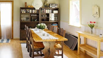 A dining area with a wooden table, chairs, a chandelier, bookshelves, and a window with flowers.