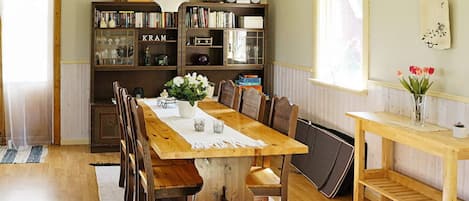 Un comedor con mesa de madera, sillas, candelabro, estanterías y una ventana con flores.