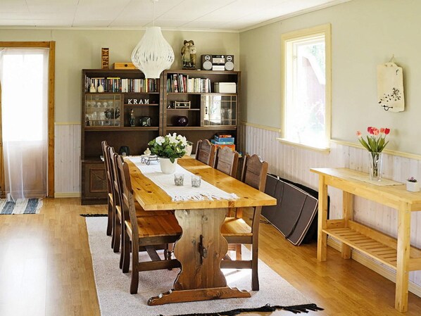 A dining area with a wooden table, chairs, a chandelier, bookshelves, and a window with flowers.