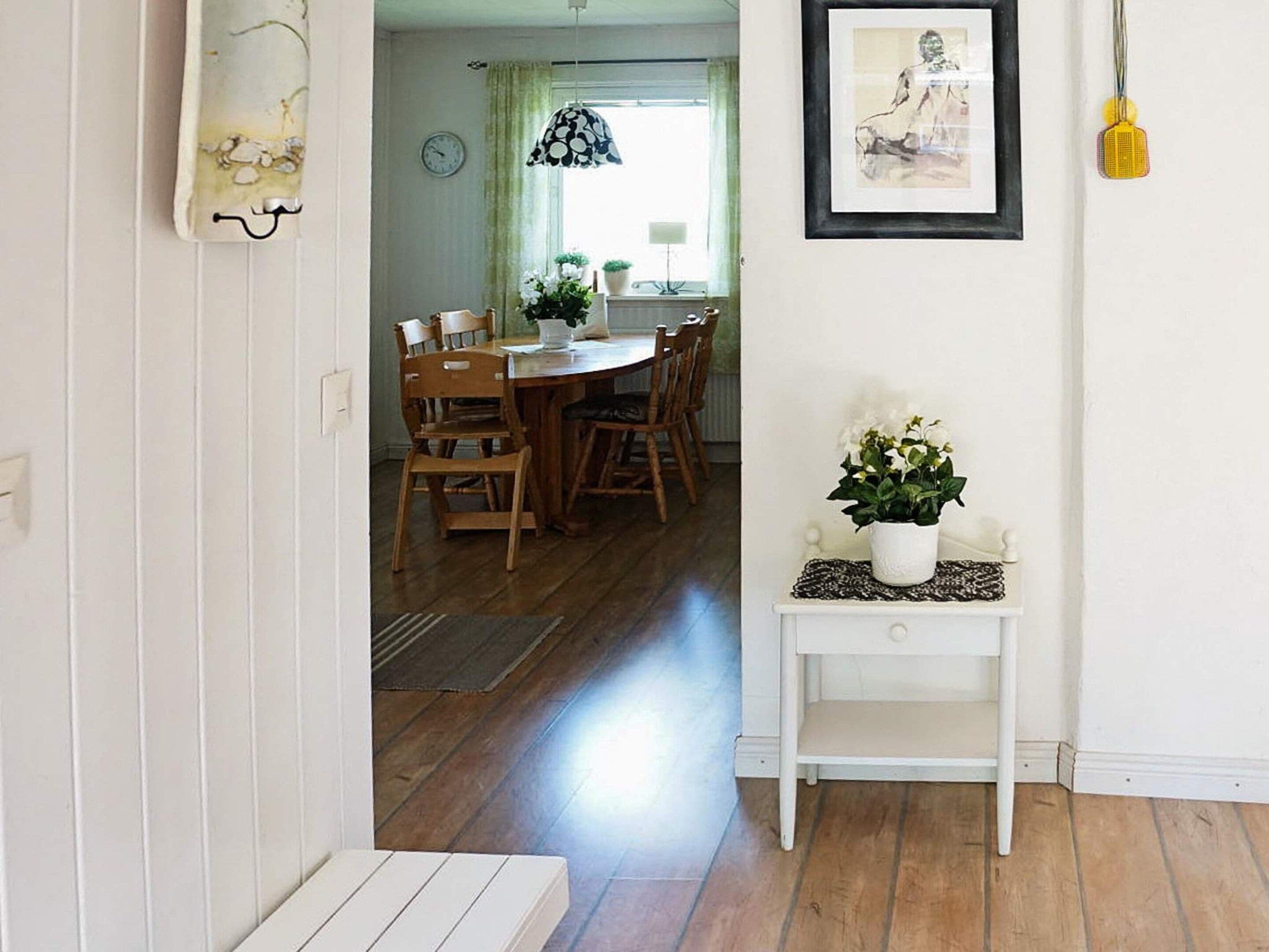 A hallway leading to a dining area with wooden chairs and a table, a white console table with a plant, and a framed artwork on the wall.