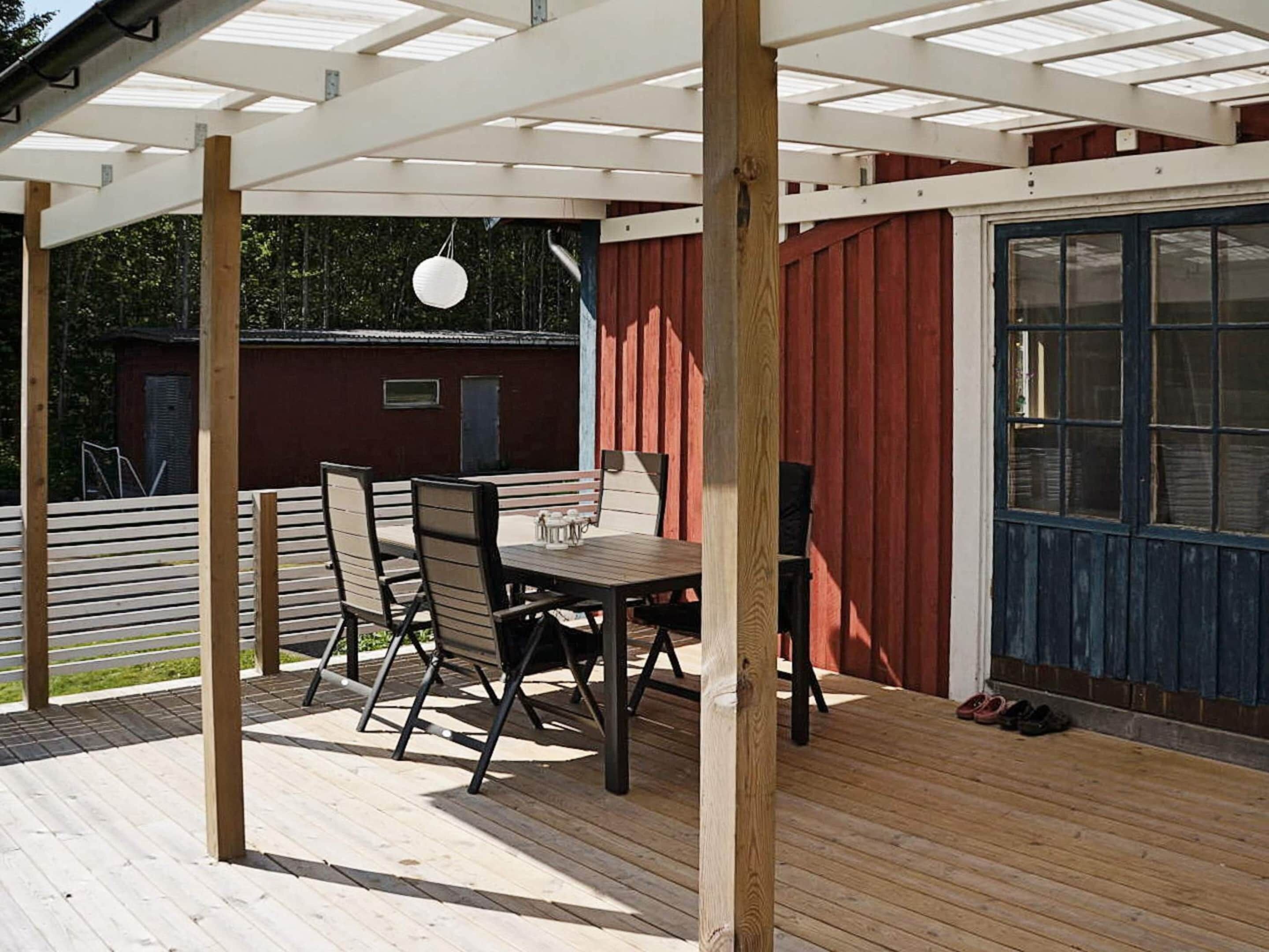 An outdoor patio with a wooden table and chairs, a pergola with a hanging light, and a red building in the background.