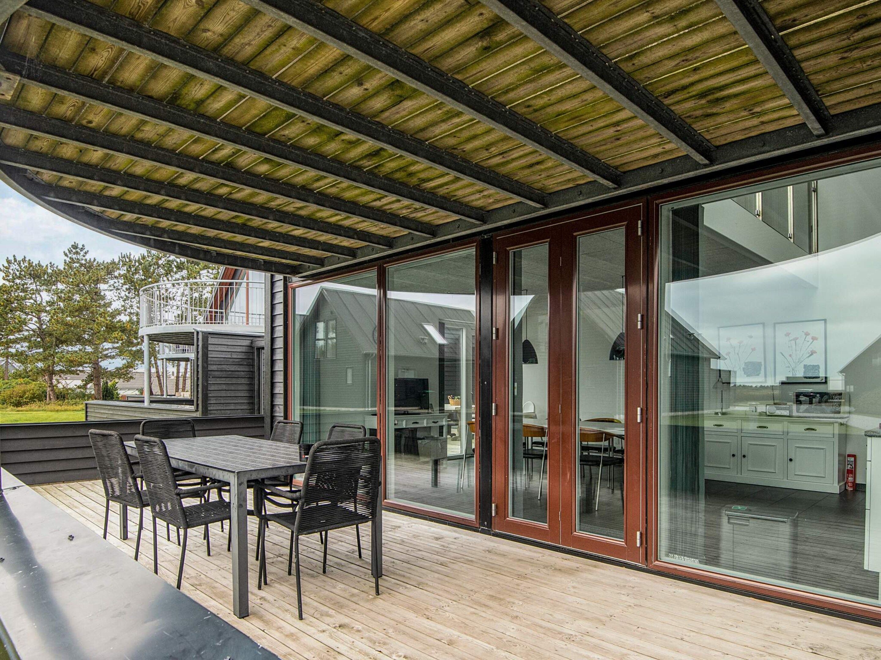 Ein überdachter Außenbereich mit Tisch und Stühlen, große Glasüren zu einer Küche und eine Holzterrasse.