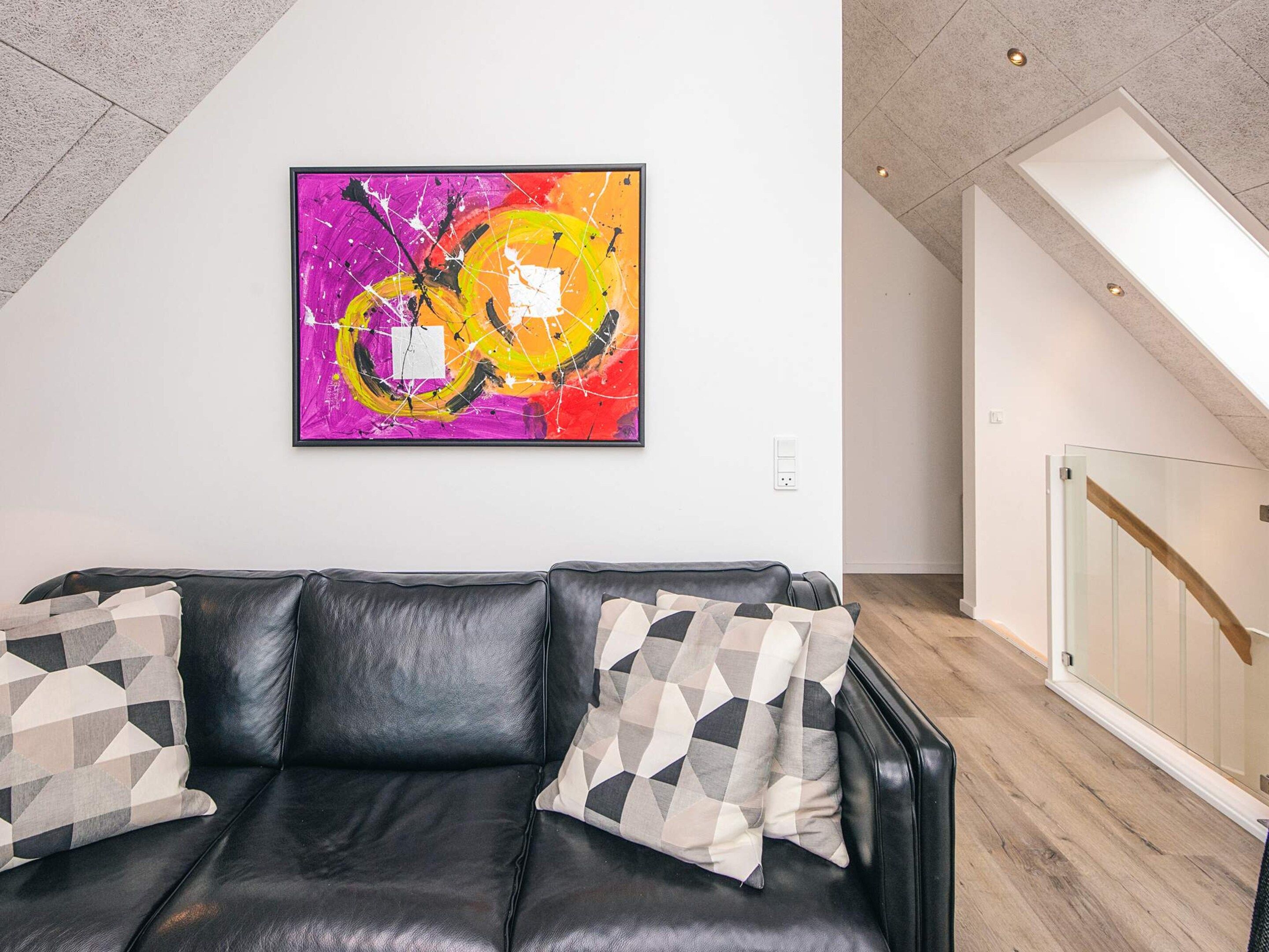 A modern living room with a black leather sofa, geometric patterned cushions, and a vibrant abstract painting on the wall.