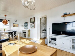 Un salon moderne doté d’un grand téléviseur à écran plat, d’un parquet, d’un coin repas avec une table et des chaises, et d’un meuble blanc avec des étagères.