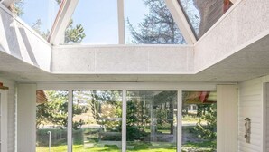 A sunlit room with a large glass door leading to a grassy yard, a table with chairs, and a window with a view of trees. - 4 Star Holiday Home in Blavand (Blavand)