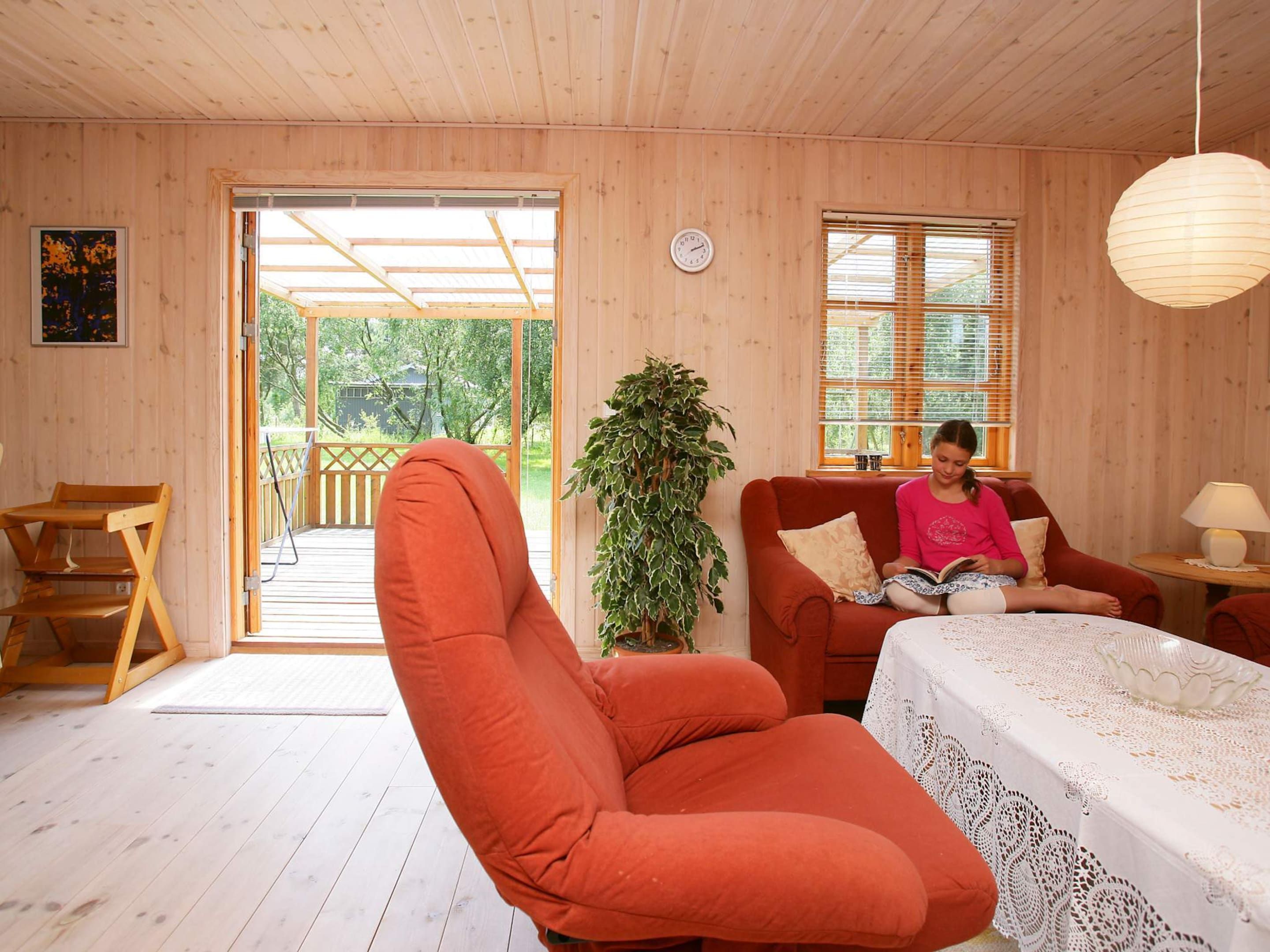 Une pièce chaleureuse avec un décor intérieur en bois, un fauteuil rouge, un canapé, une table recouverte d’une nappe en dentelle et une plante en pot.