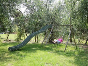A playground with a slide and swings in a grassy area surrounded by trees.