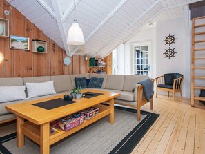 Uma sala de estar aconchegante com teto de madeira, um sofá, uma mesa de centro com jogos de tabuleiro e uma estante de livros.