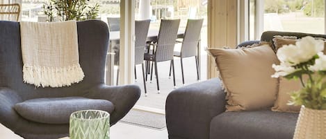 A modern living room with a grey sofa, a matching armchair, and a dining area with wicker chairs.