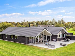 Et moderne hus med en stor terrasse, et overdækket udendørs siddeområde og udsigt over det omkringliggende landskab.