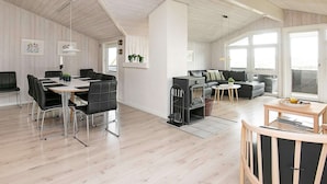 A spacious living area with a high ceiling, a dining table with chairs, a sofa, a fireplace, and large windows allowing natural light.