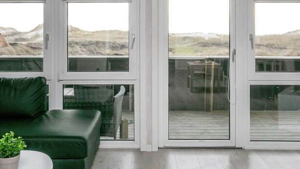 A room with a green sofa, a small table, and a potted plant. There are large glass doors leading to an outdoor area with a view of a grassy landscape.