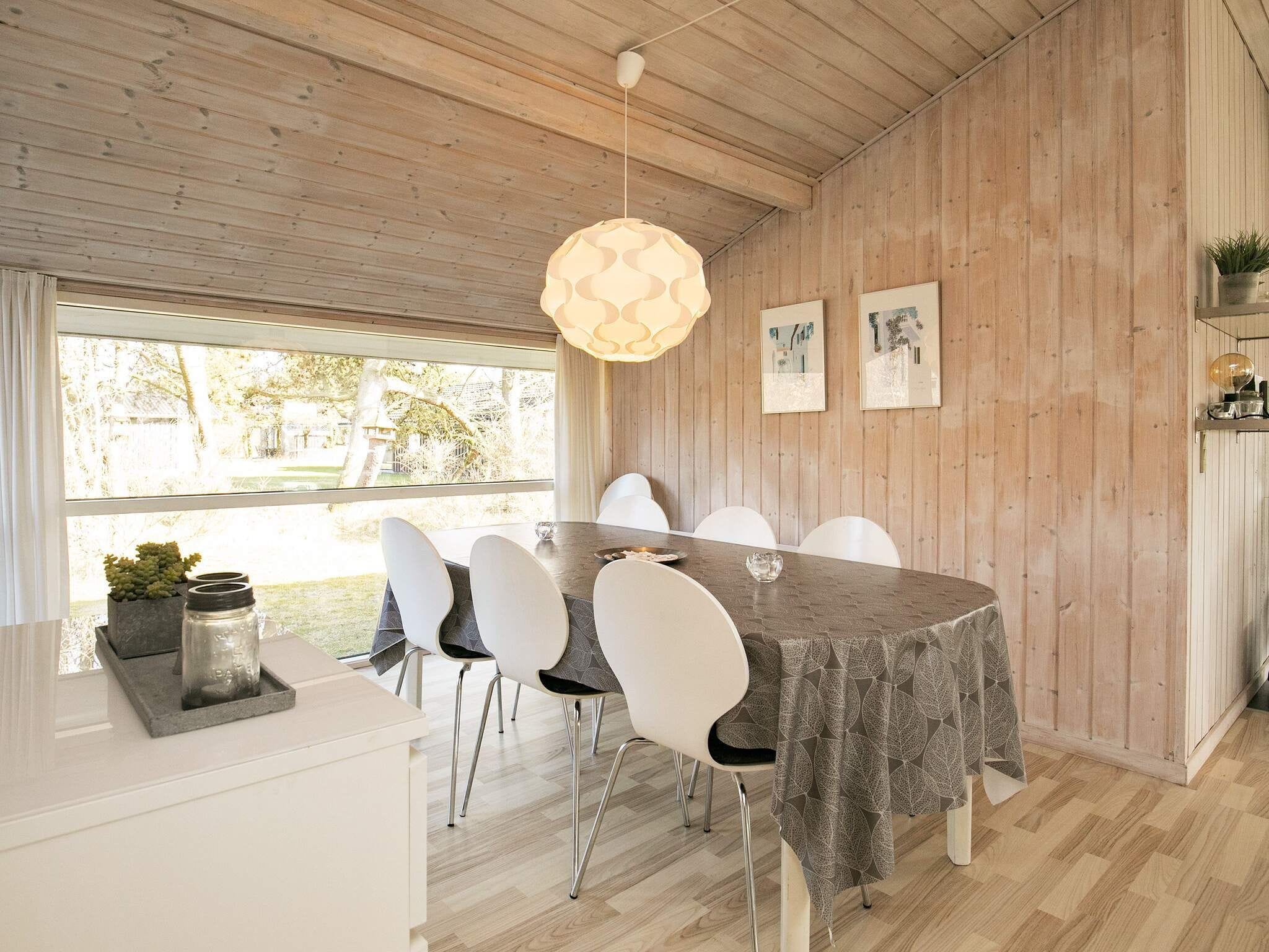 A dining area with a wooden ceiling, a table with a leaf-patterned tablecloth, white chairs, and a hanging light fixture.
