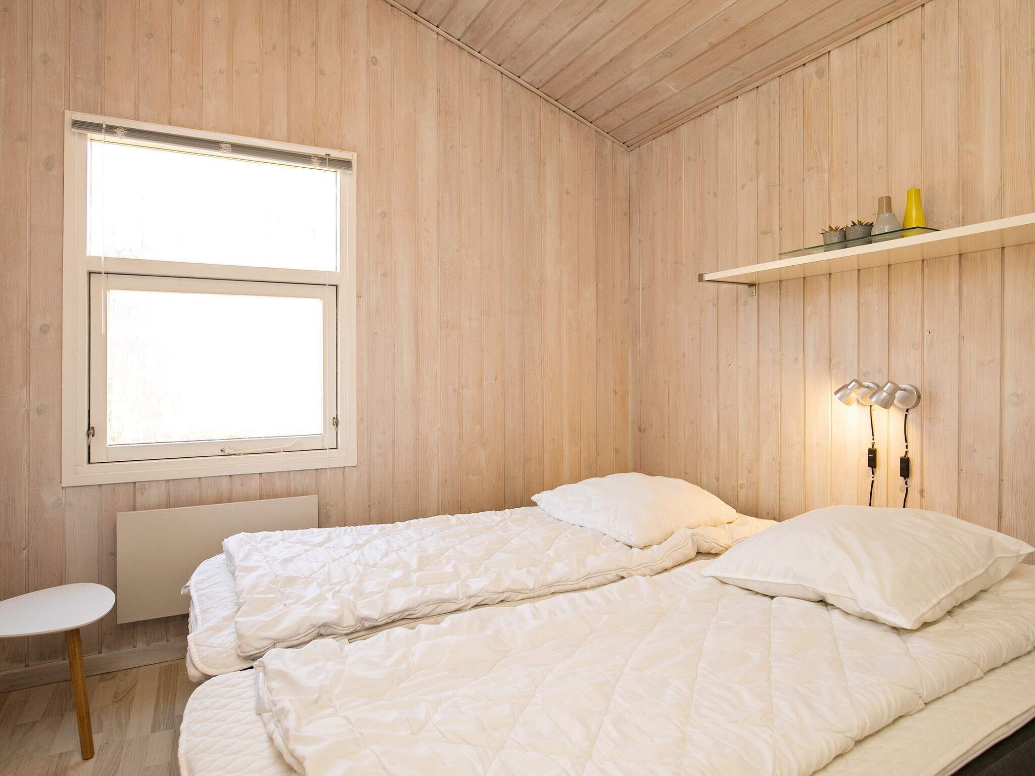 A bedroom with a bed, a window, a bedside table, and a shelf with decorative items.