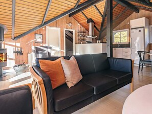 L’intérieur d’un chalet accueillant, avec un plafond en bois, un canapé noir agrémenté de coussins à motifs, un coin repas avec une table et des chaises, et une cuisine équipée d’appareils électroménagers blancs.