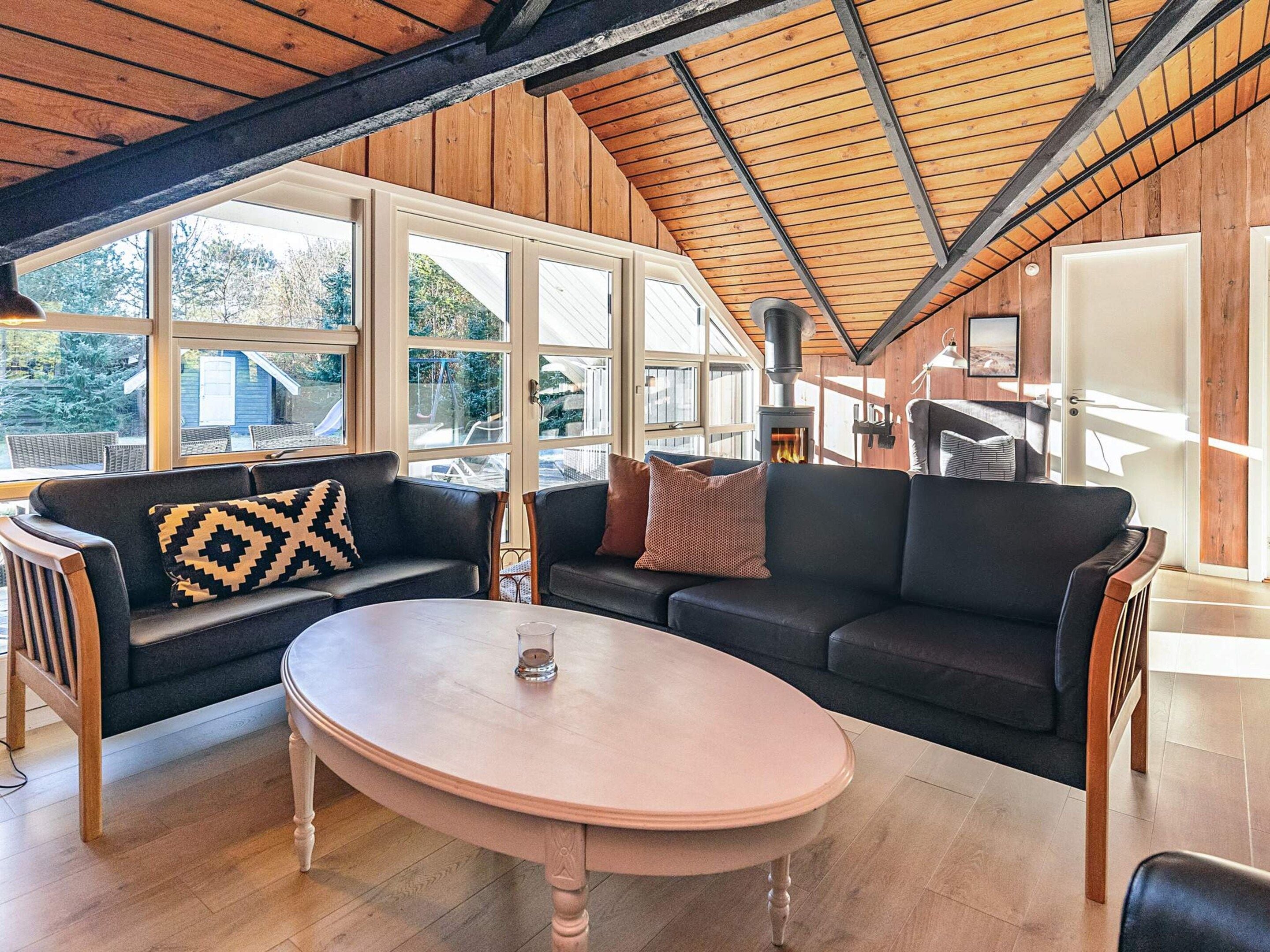 A cozy living room with a wooden ceiling, large windows, a black sofa, a round table, and a fireplace.