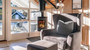 A cozy room with a large window, a comfortable armchair, a small ottoman, and a wood-burning stove.