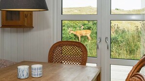 A dining area with a wooden table, two patterned cups, a wicker chair, and a view of a deer through a glass door. - Modern Holiday Home With Large Terrace in Fanø (Fano)