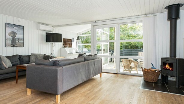 A modern living room with a grey sofa, wooden flooring, a black wood-burning stove, and a large glass door leading to an outdoor area. - Modern Holiday Home With Large Terrace in Fanø (Fano)