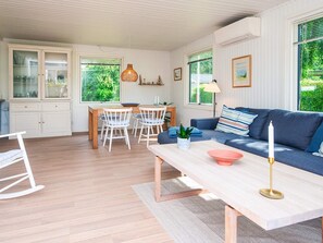 En moderne stue med sofa, spisebord med stole, en lænestol og et stort vindue med udsigt til grøn vegetation.