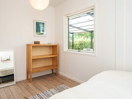 Un dormitorio con cama, repisa de madera, un cuadro enmarcado y ventana con vista a follaje.