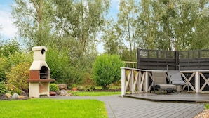 A patio with a stone barbecue grill, outdoor seating, and a wooden deck. - Holiday Home in Otterndorf (Otterndorf)