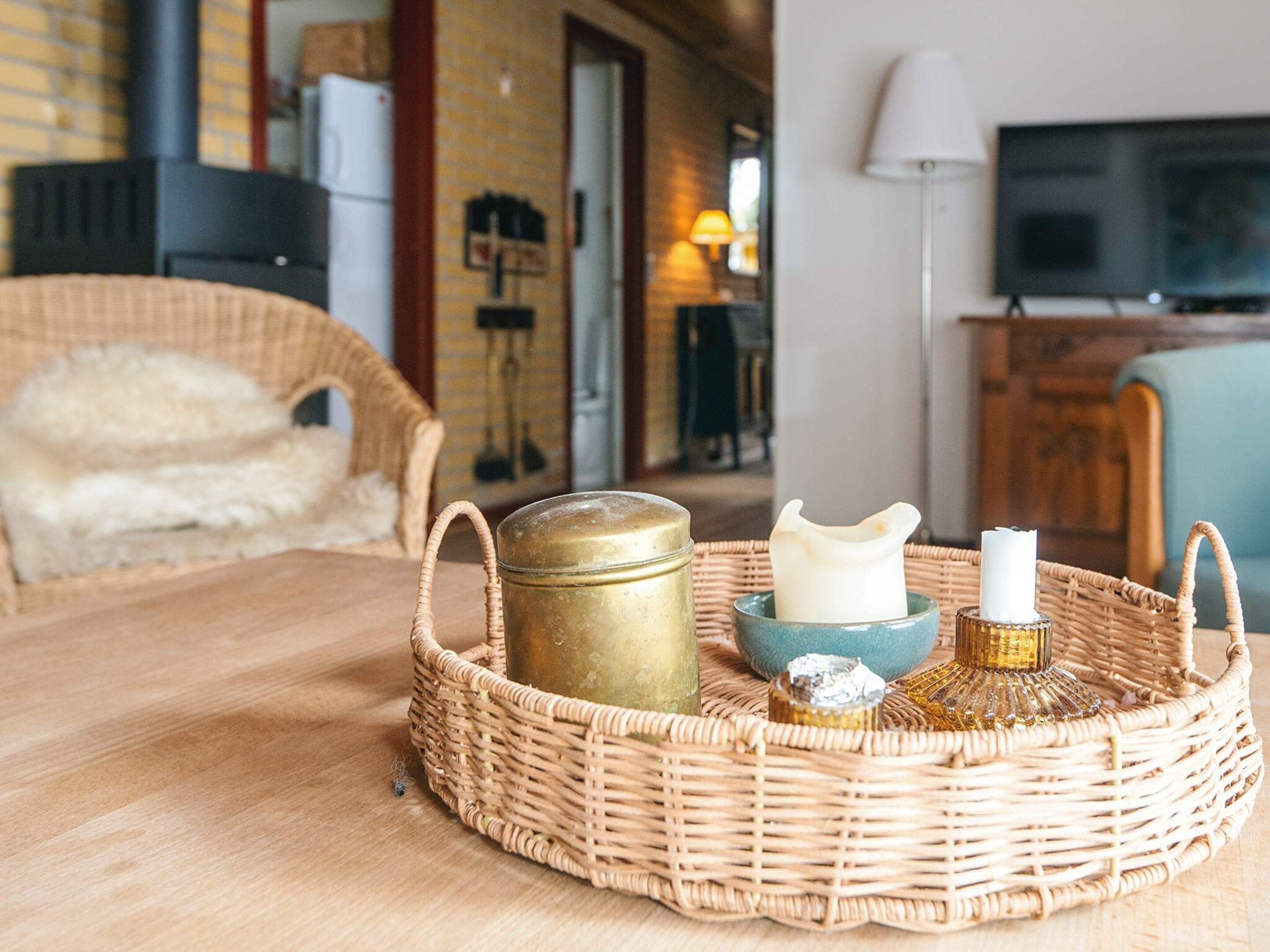 A wicker tray with a jar, candle, and decorative item on a wooden table in a room with a TV and a lamp.