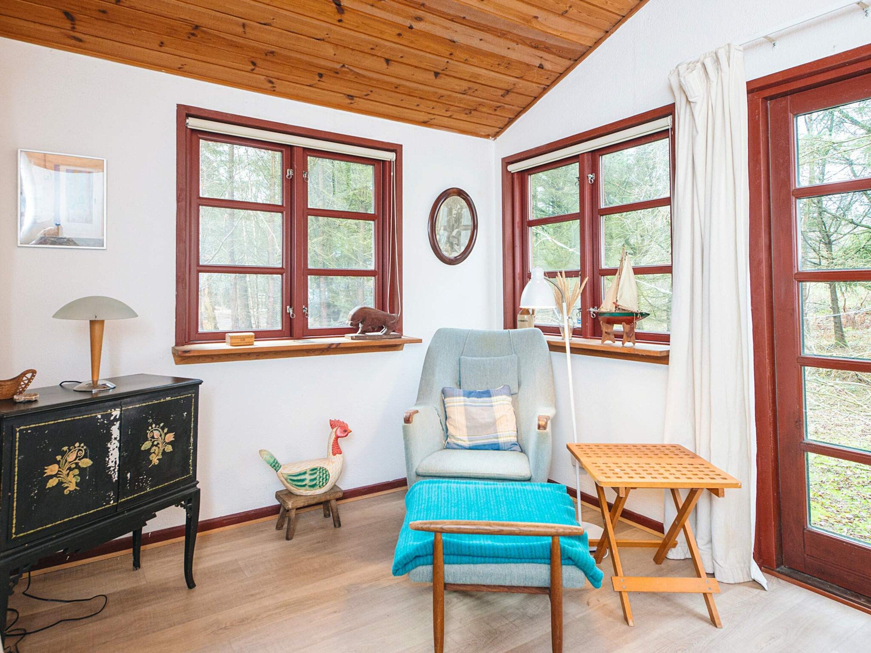 A room with a wooden ceiling, a blue chair, a small table, a wooden cabinet, and two windows with curtains.