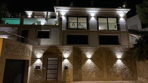 Una casa moderna de dos pisos con fachada de piedra, una entrada central con portón metálico y numerosas ventanas iluminadas.