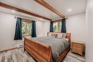 A bedroom with a wooden bed, two windows with curtains, a bedside table, and a radiator.