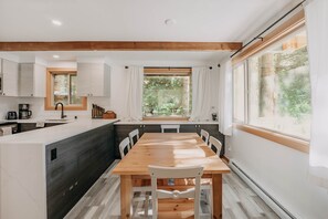 A modern kitchen with a wooden dining table, white cabinets, and large windows.