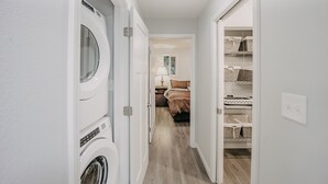 Une buanderie avec des lave-linge et des sèche-linge superposés, une chambre avec un lit et une table de chevet, et un espace de rangement avec des paniers.