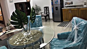 Un salon comprenant une table en verre, des chaises, un canapé, un réfrigérateur et un coin cuisine.