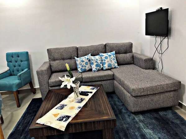 Un salon comprenant un canapé d’angle, un fauteuil bleu, une table basse avec un set de table, et un téléviseur à écran plat fixé au mur.