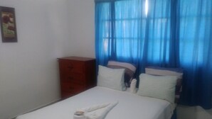 A neatly made bed with a towel and a cup, a wooden dresser, and blue curtains. - Room-room With fan use Bavaro Punta Cana (Punta Cana)