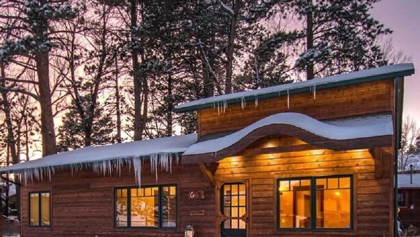 Uma cabana de madeira com telhado coberto de neve, janelas amplas e luz interior aconchegante.