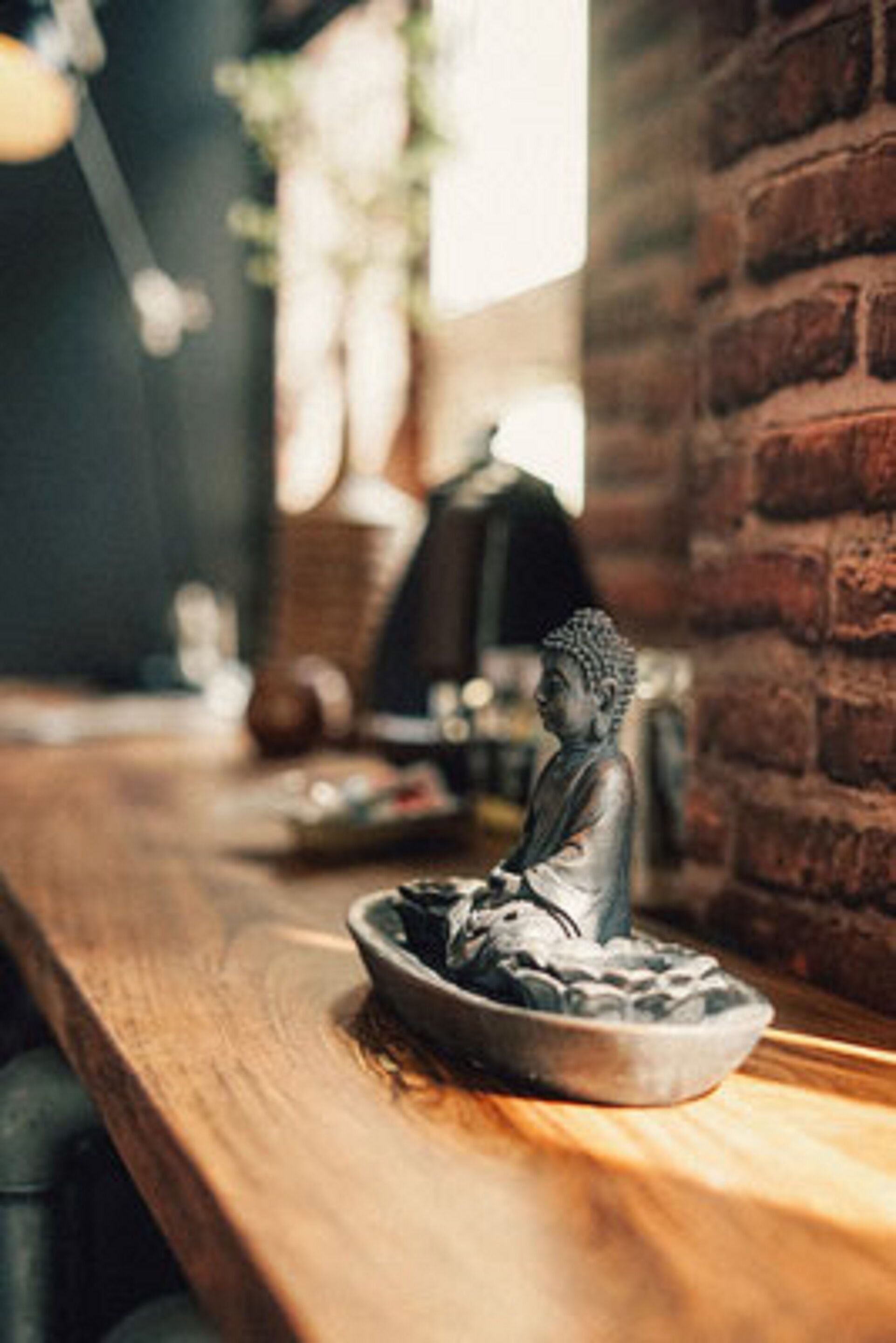 A Buddha statue on a tray on a wooden table in a room with brick walls and a window.