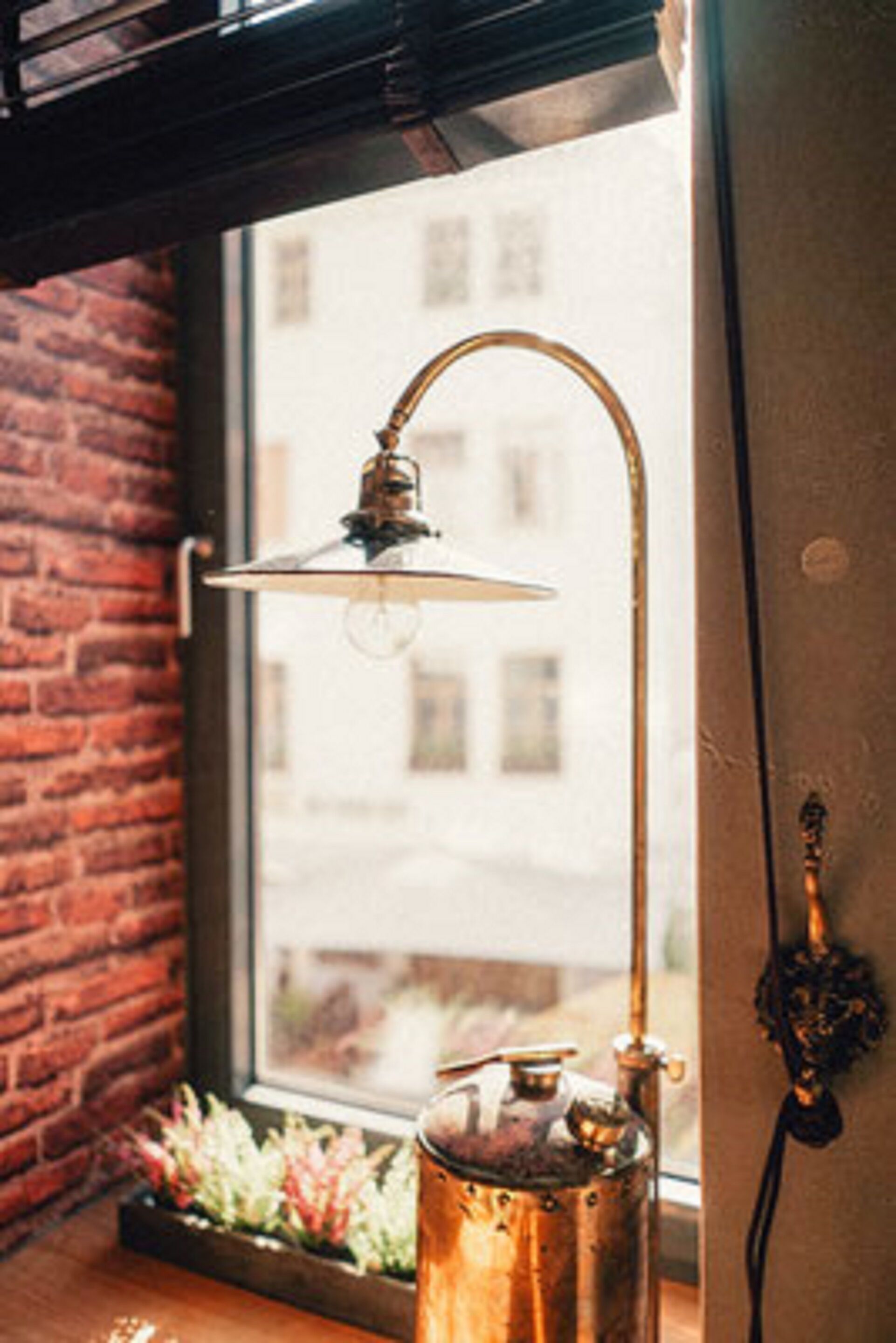 A rustic interior with a brick wall, a window with a view of buildings, a brass lamp, and a copper pot on a windowsill.
