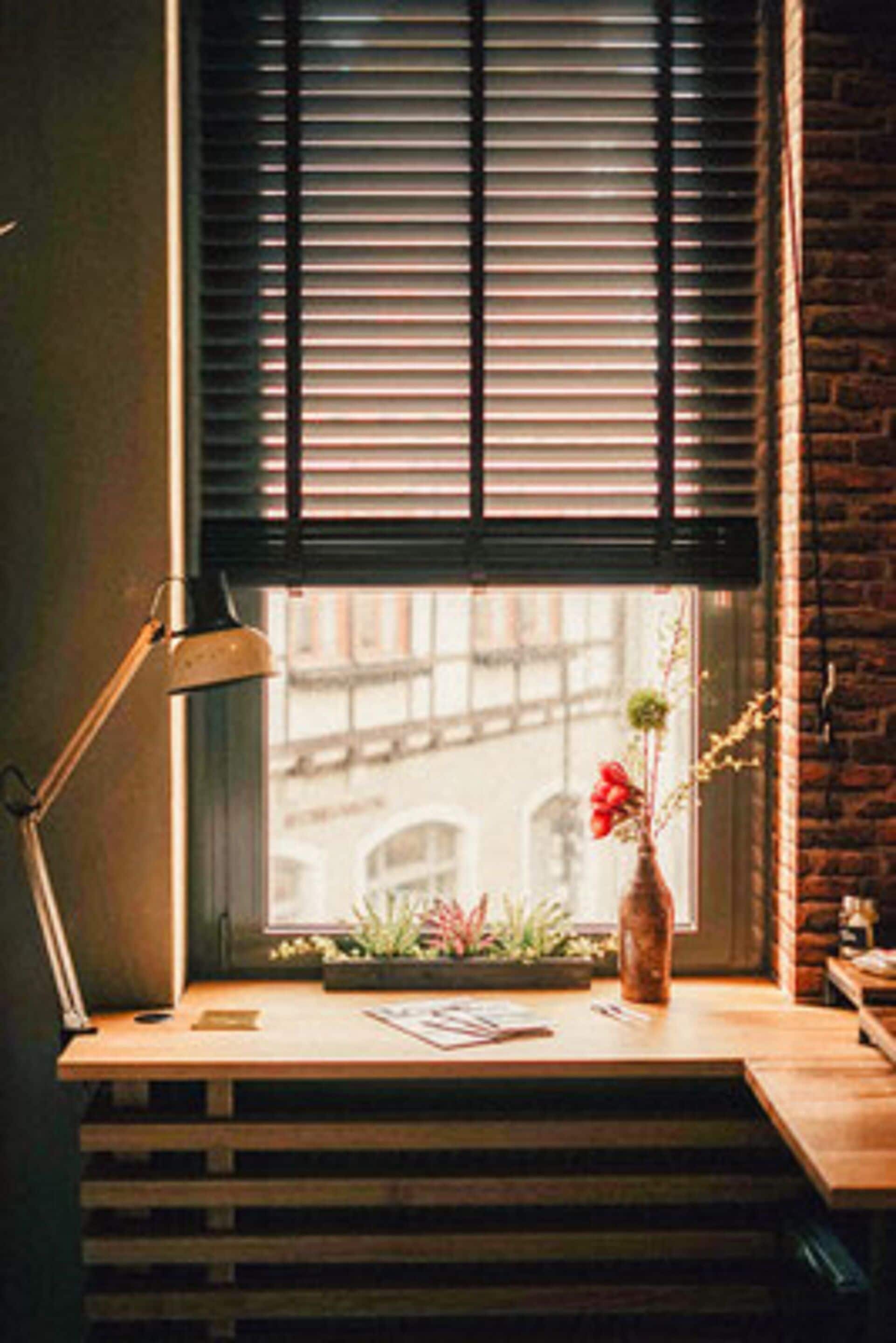 A desk with a lamp, a vase with flowers, and a window with blinds.