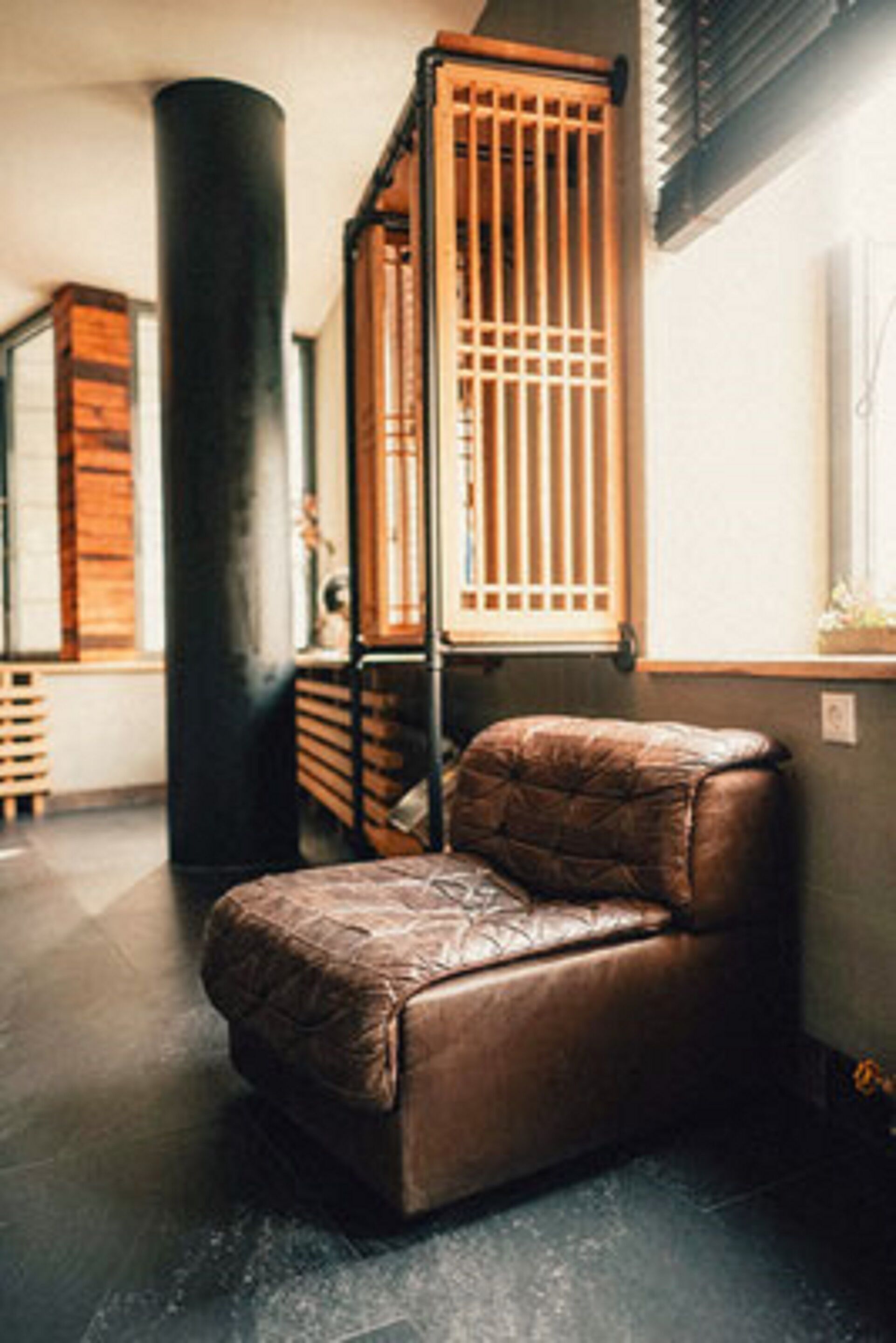 A brown leather chair in a room with a large black column, wooden slats, and a window with blinds.